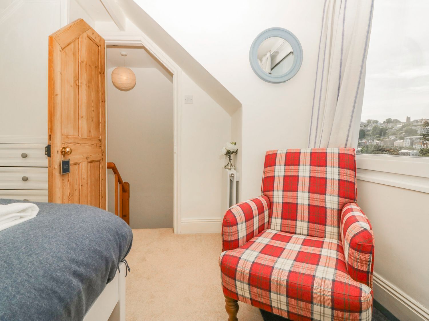 A bedroom with a bed, a plaid armchair, a wooden door, and a view of a city through a window.