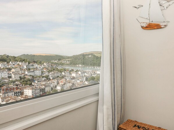 Une pièce avec vue sur une ville côtière, une fenêtre ornée de rideaux blancs et une petite décoration représentant un navire accrochée au mur.