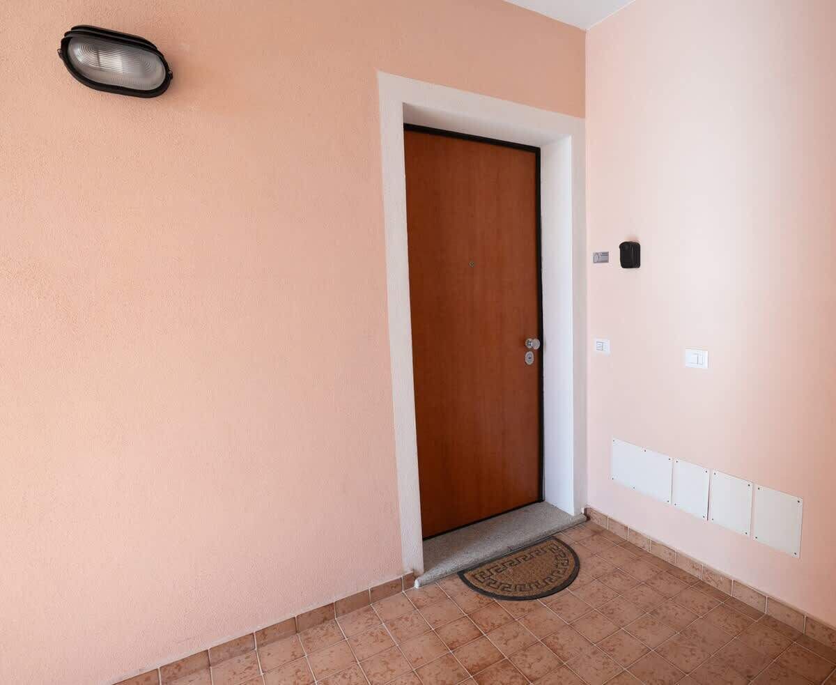 Um corredor com porta de madeira, tapete de entrada e luminária na parede.