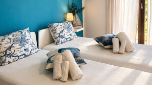 Habitación de hotel con dos camas, una pared azul, una ventana con cortinas y una planta pequeña en una mesita de noche.