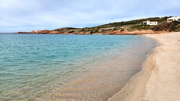 Una playa con agua cristalina, orilla de arena y casas al fondo.