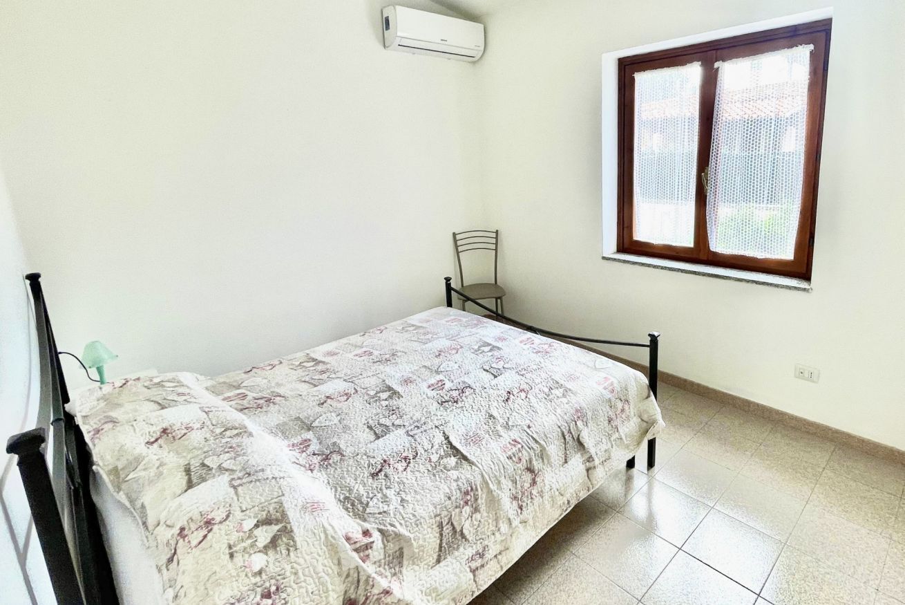 Un llit individual amb un edredó floral en una habitació amb finestra, una cadira i un aparell d'aire condicionat.