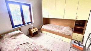 A bedroom with two single beds, a wooden wardrobe, a small bedside table with a lamp, and a window with curtains.
