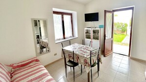 A small, well-lit room with a dining table, chairs, a sofa, a TV, and a door leading to a garden.