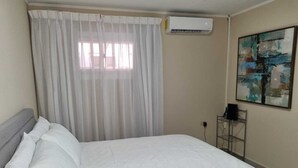 A neatly made bed with white linens, a window with sheer curtains, an air conditioning unit on the wall, and a metal shelf with a speaker. - Taminaka Comfortable III (Oranjestad)