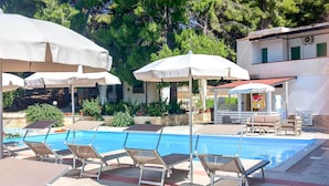 Ein Poolbereich mit Liegestühlen und Sonnenschirmen, umgeben von Grünflächen und einem Gebäude mit rotem Dach.