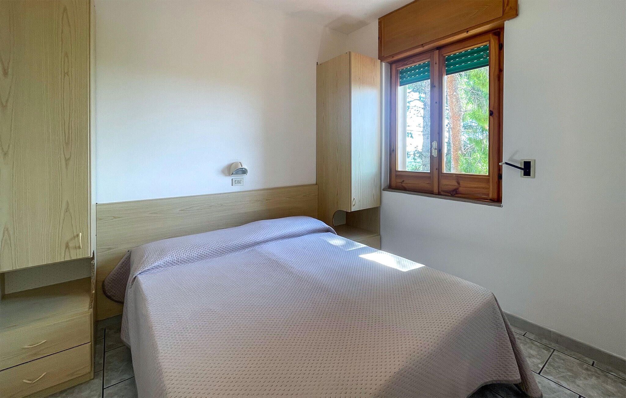 A small, single-bed room with a wooden wardrobe, a window with blinds, and a wall-mounted light.