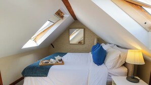 A bedroom with a sloped ceiling, a bed with white bedding, a blue pillow, a bedside table with a lamp, and a tray with tea items. - Parrog Bach (Newport)