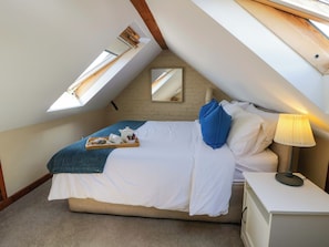 A bedroom with a sloped ceiling, a bed with white bedding, a blue pillow, a bedside table with a lamp, and a tray with tea items.