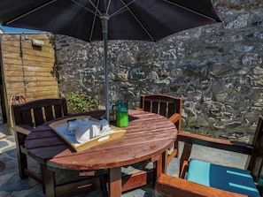 A round wooden table with chairs, a table umbrella, and a stone wall in the background.