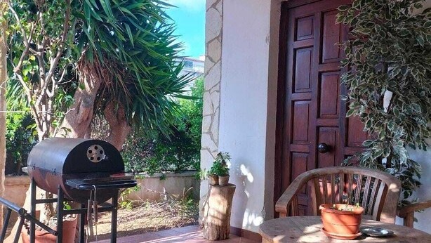 Een terras buiten met een barbecue, een houten tafel en plantenpotten.