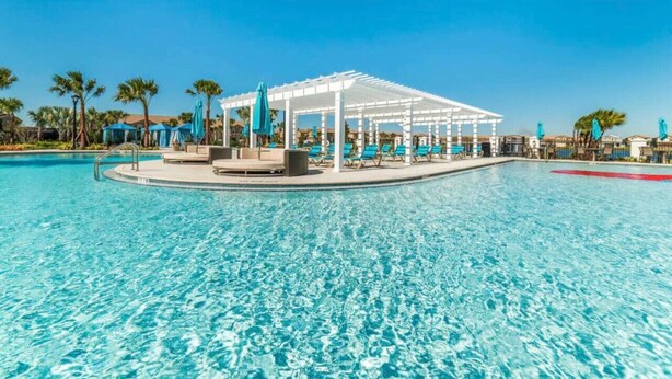A swimming pool with a covered seating area, lounge chairs, and palm trees in the background. - 5 Bedroom 4 Bathroom Windsor at Westside 8933PW (Kissimmee)