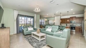 A spacious living room with a sofa set, a coffee table, and a chandelier. There is a kitchen area with wooden cabinets and a refrigerator. - 5 Bedroom 4 Bathroom Windsor at Westside 8933PW (Kissimmee)