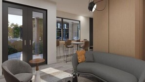 A modern living room with a grey sofa, a small round table, and a chair. There is a large window with a view of the outside.