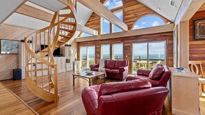 Una habitación espaciosa con una escalera espiral de madera, ventanas amplias con vistas al océano y una zona de estar acogedora con sofás de cuero rojo.