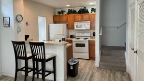 A compact kitchen with wooden cabinets, a white refrigerator, a microwave, and a stove. There are two chairs at a small island, and a trash bin is visible. - 4 Bedroom 3 Bathroom Villas At Seven Dwarfs 8-107 (Kissimmee)