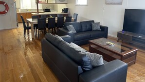 Ett modernt vardagsrum med en soffa, ett soffbord och en tv. Det finns en matplats med stolar och ett bord, och ett kök syns i bakgrunden.