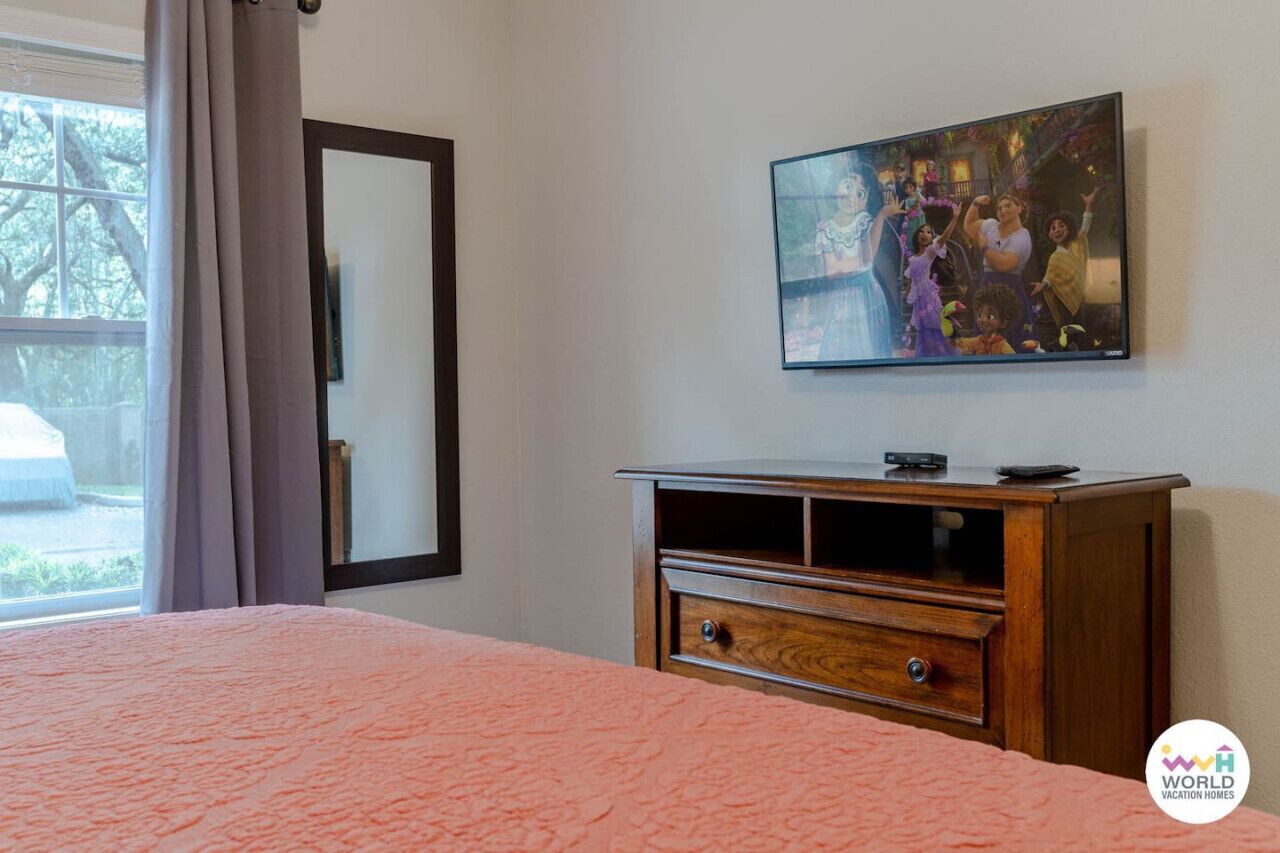Une chambre à coucher avec un lit, une commode en bois, un téléviseur fixé au mur et une fenêtre avec des rideaux.