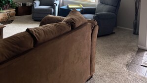 Ein Wohnzimmer mit zwei Sesseln, einem Sofa, einer Lampe, einem Gemälde und einem Fenster mit Vorhängen.
