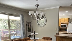 Ein Essbereich mit Holztisch und braunen Stühlen, einer Chandelier, einer Wanduhr und Blick auf eine Terrasse mit Bank und Topfpflanze.