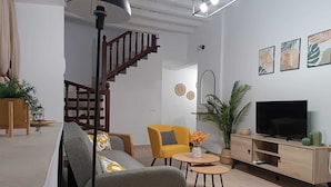 A modern living room with a staircase, a sofa, a yellow armchair, a TV, and decorative plants.