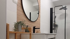 A modern bathroom with a wooden vanity, a round mirror, a shower with a black frame, and neatly rolled towels on a wooden rack.