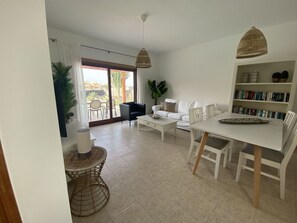 Relax in this open-plan living space with a dining area, sofa, and bookshelf, leading to a patio with outdoor seating.