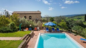 A swimming pool with lounge chairs and umbrellas, surrounded by greenery and a stone building.