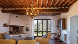 A cozy living room with a fireplace, wooden beams, and a large window with a view of trees.