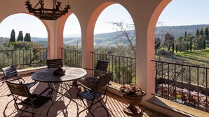 A terrace with a round table and chairs, a chandelier, and a view of the countryside through arches.