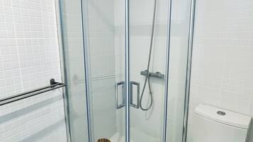 A glass-enclosed shower with a handheld showerhead, a toilet, and a trash bin.