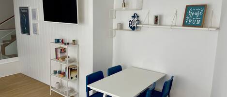 Een moderne kamer met een witte tafel en blauwe stoelen, een boekenkast, een wandtelevisie en decoratieve objecten op planken.