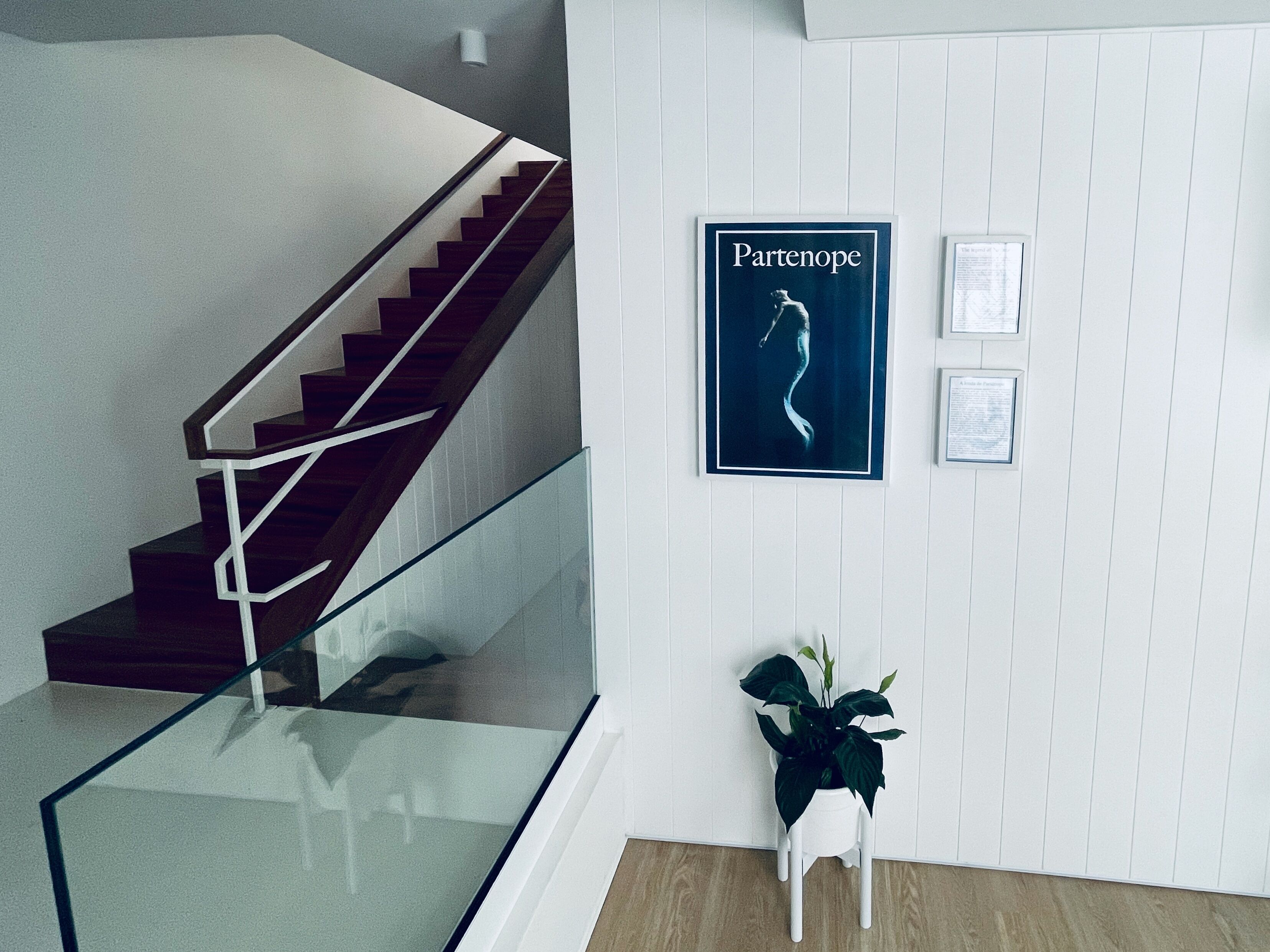 Una escalera con barandillas de vidrio, una obra de arte enmarco titulada 'Parthenope', y una planta en maceta sobre una silla blanca.