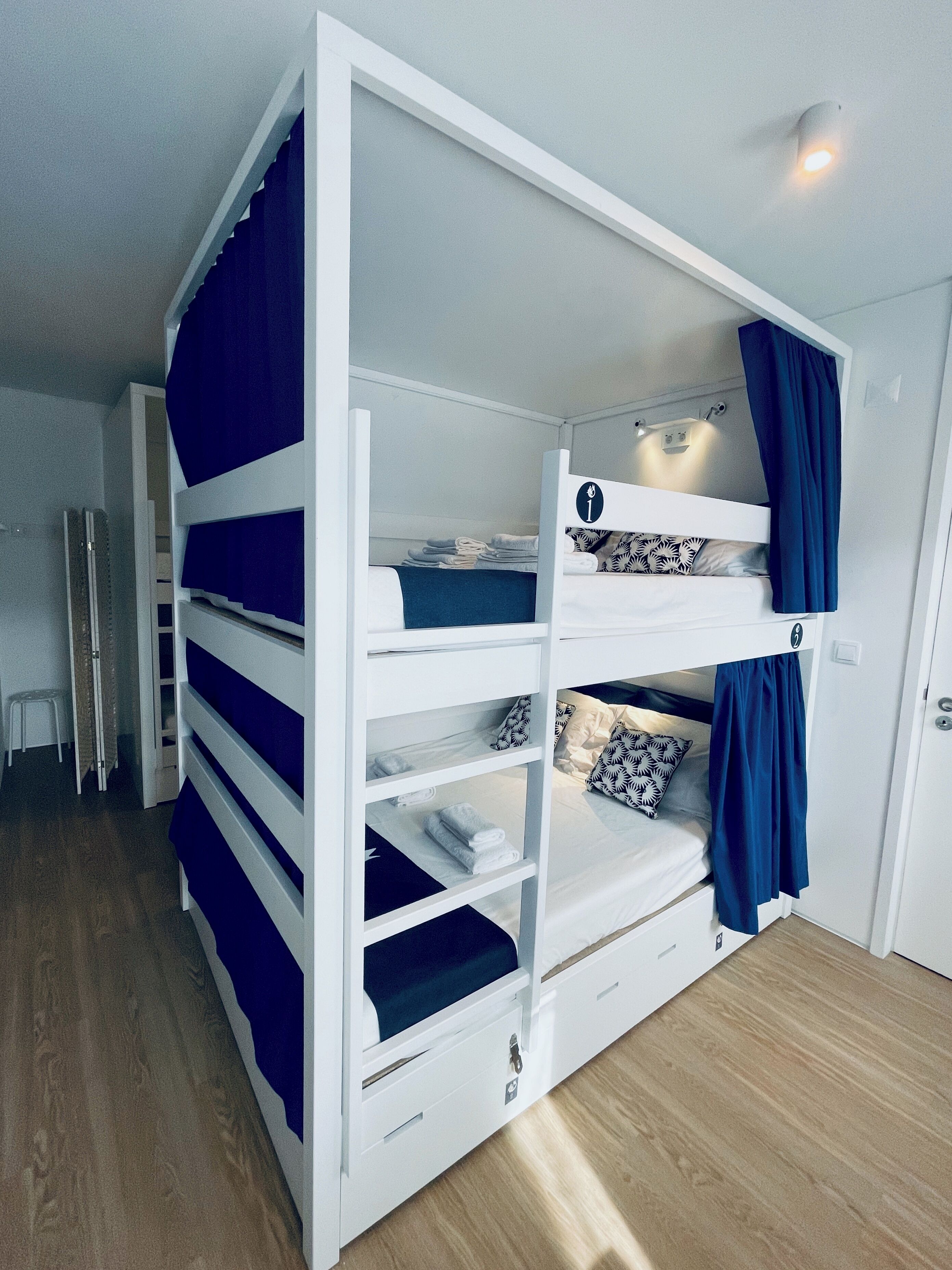 A bunk bed with a white frame and blue curtains, a small stool, and a door in the background.