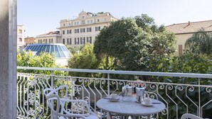 En balkon med hvide metalstole og et bord, med udsigt over et bybillede med bygninger og træer.