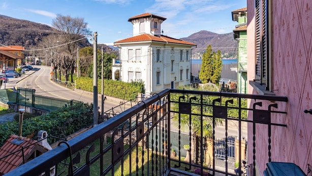 A balcony with a view of a white building with a red roof, a road, and mountains in the background. - Wonderful  villa for 8 guests with WIFI, TV and balcony (Porto Ceresio)