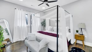 Una cama con dosel y cuatro columnas, ropa de cama blanca y un plaid rojo, una mesita de noche con lámpara y una planta pequeña.