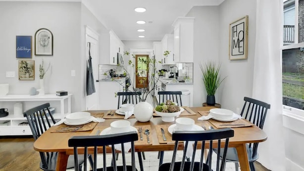 Un comedor con mesa de madera, sillas blancas y un jarrón central con follaje.
