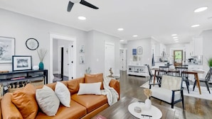 Una sala de estar moderna con un sofá de cuero marrón, un comedor con una mesa y sillas de madera, y una cocina al fondo.