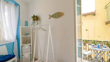 Una stanza con una sedia bianca, uno scaffale bianco con cesti, uno stendibiancheria e un balcone con tavolo e sedie.