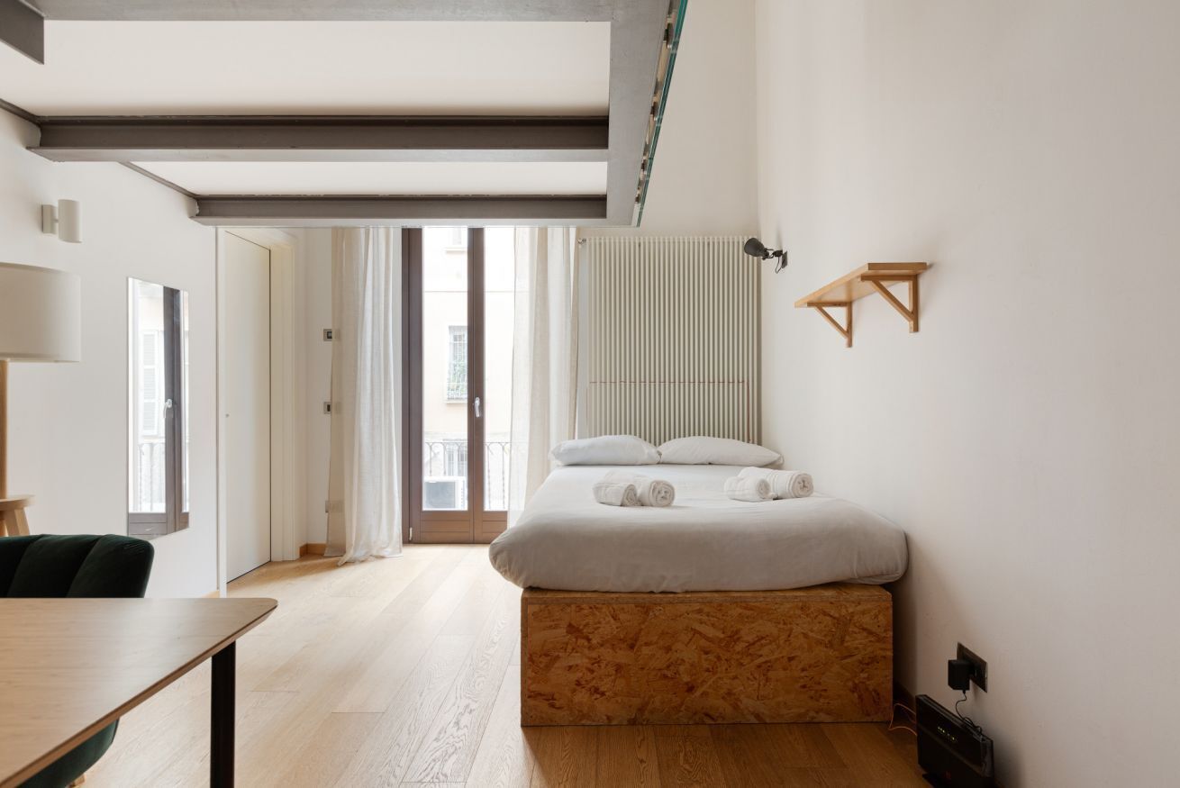 Ein modernes Hotelzimmer mit einem Bett mit Holzrahmen, einem Schreibtisch, einem Stuhl, einem Heizkörper und Blick auf die Stadt durch das Fenster.