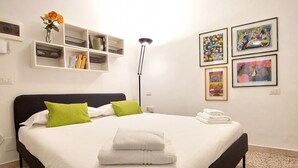 A neatly made bed with white linens and green pillows, a bedside table with a lamp, and artwork on the wall.