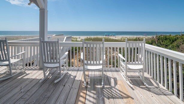Eine Holzterrasse mit weißen Geländern und Stühlen mit Blick auf einen Strand.