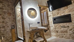 Un baño rústico con pared de piedra, vigas de madera, un espejo redondo, un lavabo y un televisor montado en la pared.