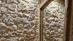 Una habitación con pared de piedra, suelo de madera y un espejo que refleja la pared. Hay dos jarrones verdes con plumas blancas y una lámpara geométrica negra sobre un soporte de madera.