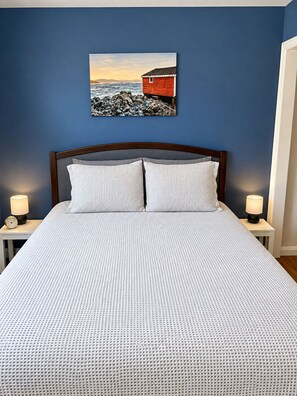 A bedroom with a bed, two bedside tables, a nightstand, and a wall-mounted picture of a red building by the sea. - Cozy 2 bedroom bungalow overlooking scenic Twillingate harbour (Twillingate)