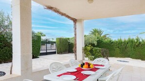A covered outdoor dining area with a table set for six, surrounded by white chairs, and a view of a garden and gate in the background.
