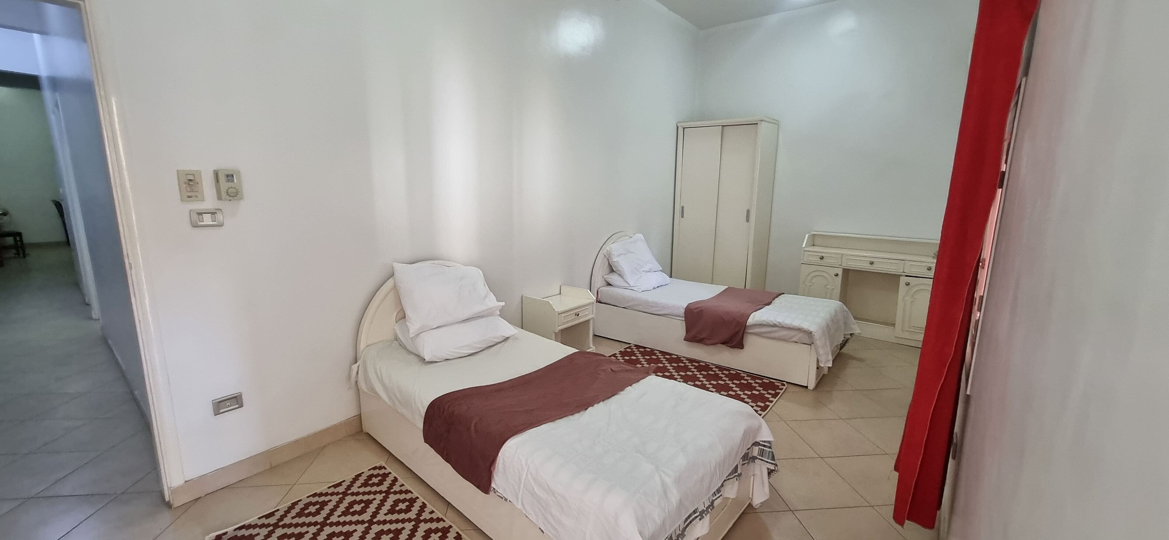 A bedroom with two beds, a wardrobe, a desk, and a patterned rug.