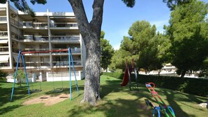 A playground with swings and a slide, surrounded by trees and a building in the background. - Beautiful apartment for 6 people with WIFI, pool, A/C, TV, terrace and pets allowed (Salou)