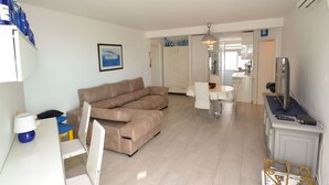 A modern living room with a beige sectional sofa, a dining table with chairs, a TV on a stand, and a kitchen area in the background.